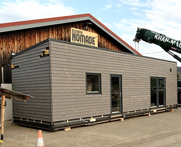 Kleinernomade-modulhouse-modell10-tinyhouse-370x300 Modulares Tiny House mit dunkelgrauer Holzfassade vor einer Werkhalle mit "Kleiner Nomade"-Schild.