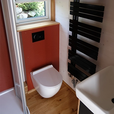 Kleinernomade-modulhouse-modell10-tinyhouse-380x380-4 Kompaktes Badezimmer in einem Tiny House mit wandhängendem WC, Fenster, modernem Heizkörper und Holzfußboden.