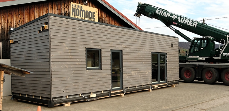 Kleinernomade-modulhouse-modell10-tinyhouse-780x380-3 Modulares Tiny House mit dunkelgrauer Holzfassade vor einer Werkhalle mit "Kleiner Nomade"-Schild.