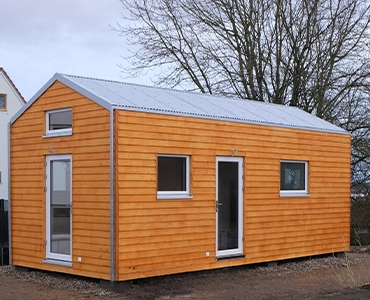 Kleinernomade-modulhouse-modell9-tinyhouse-370x300 Modulares Tiny House mit Holzfassade und Metall-Dach auf einem unbebauten Grundstück.