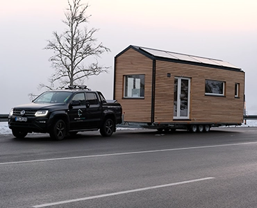 Ein Tiny House auf einem Anhänger wird von einem schwarzen Pickup auf einer verschneiten Straße transportiert.