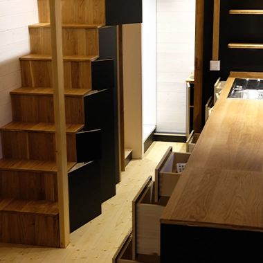 Kleinernomade-tinyhouse-modellmax72-380x380-1 Platzsparende Holztreppe mit integrierten Schubladen und Sicht auf die Küchenzeile.