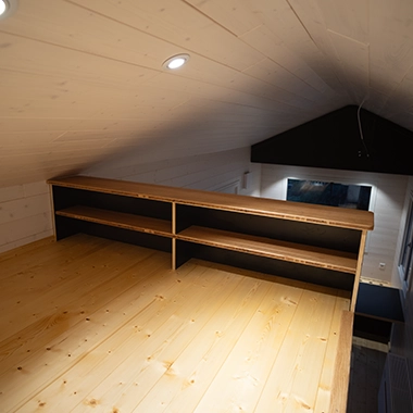 Kleinernomade-tinyhouse-modellmax72-380x380-4 Holzboden-Galerie mit eingebautem Regal und Blick auf den Wohnraum im Tiny House.