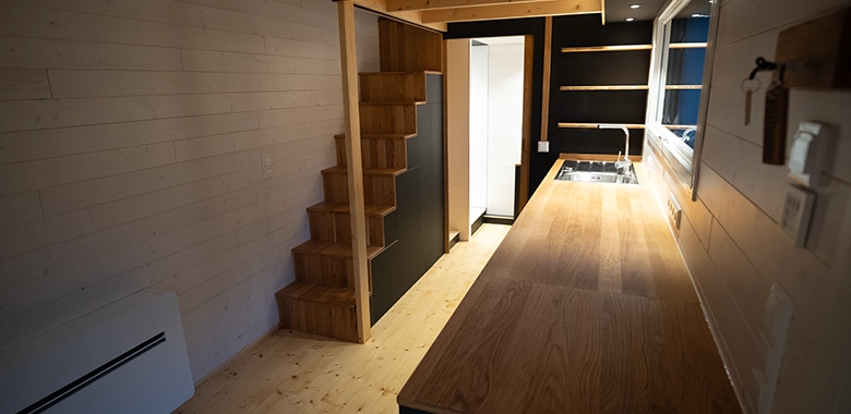 Kleinernomade-tinyhouse-modellmax72-780x380-1 Offene Wohnküche im Tiny House mit Massivholz-Arbeitsfläche und Treppe zur Galerie.