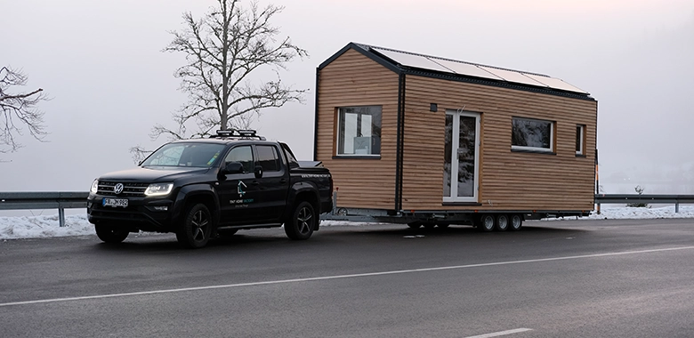 Kleinernomade-tinyhouse-modellmax72-780x380-2 Ein Tiny House auf einem Anhänger wird von einem schwarzen Pickup auf einer verschneiten Straße transportiert.