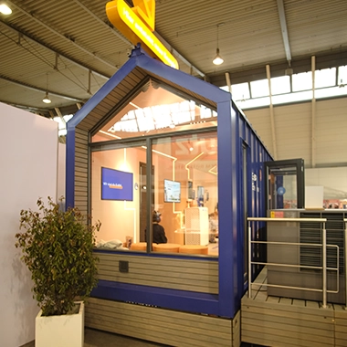 kleiner-nomade-promotion-tiny-house-enew-messe2 Blaues Tiny House mit großer Fensterfront in einer Messehalle.