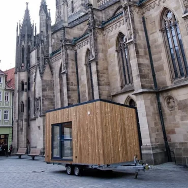 WiSi-kleiner-nomade (2) Blaues Tiny House mit großer Fensterfront in einer Messehalle.