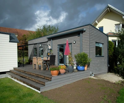 Modulhouse 10.4 ebenerdig Modernes Gartenhaus mit Holzverkleidung, Terrasse mit Tisch, Stühlen und Sonnenschirm, umgeben von Rasen und Nachbarhäusern