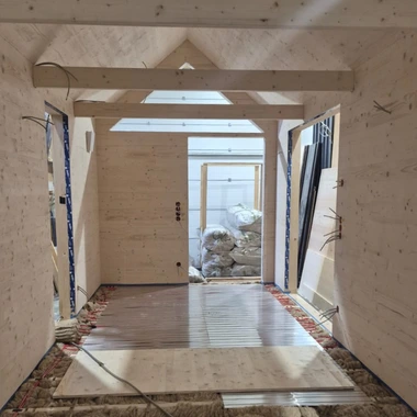 Innenraum eines Rohbaus mit Holzwänden, offenem Eingang und Baumaterialien auf dem Boden