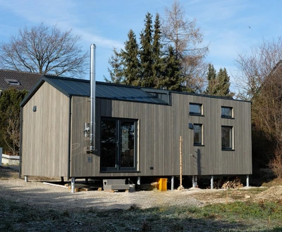 Modulhouse 10.4 mit Empore Modernes, rechteckiges Holzhaus auf Stelzen mit großen Fenstern und Schornstein in ländlicher Umgebung
