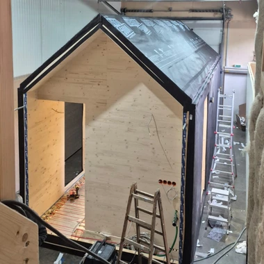 Unfertiges kleines Holzhaus mit schwarzem Dach und offenem Eingang in einer Werkstatt mit Leitern und Werkzeugen