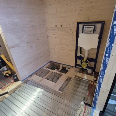 Innenraum eines Rohbaus mit Holzwänden, offenem Eingang und Baumaterialien auf dem Boden