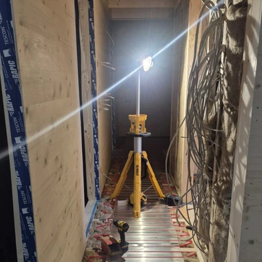 Baustelle mit einem gelben Stativlicht in einem schmalen Flur, links mit Holzplatten verkleidet, rechts mit sichtbaren Kabeln an der Wand
