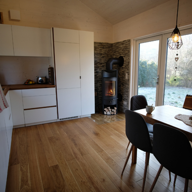 Moderner Wohnraum mit Holzboden, weißer Küchenzeile, schwarzem Kaminofen und Esstisch mit schwarzen Stühlen vor großen Fenstern