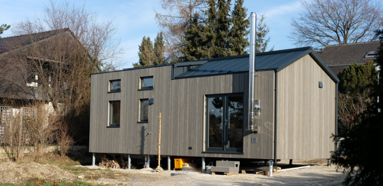 Modernes, rechteckiges Tiny House mit Holzfassade, großen Fenstern und Schornstein auf einem Grundstück mit Bäumen im Hintergrund