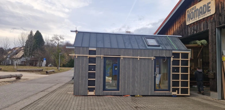 Kleines modernes Holzhaus mit großen Fenstern und Dachfenster steht auf gepflastertem Platz vor einem Gebäude mit Schild 'KLEINER NOMADE'