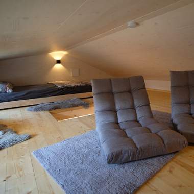 Dachzimmer mit schräger Decke, zwei gepolsterten Bodensesseln auf Teppich und niedrigem Bett im Hintergrund
